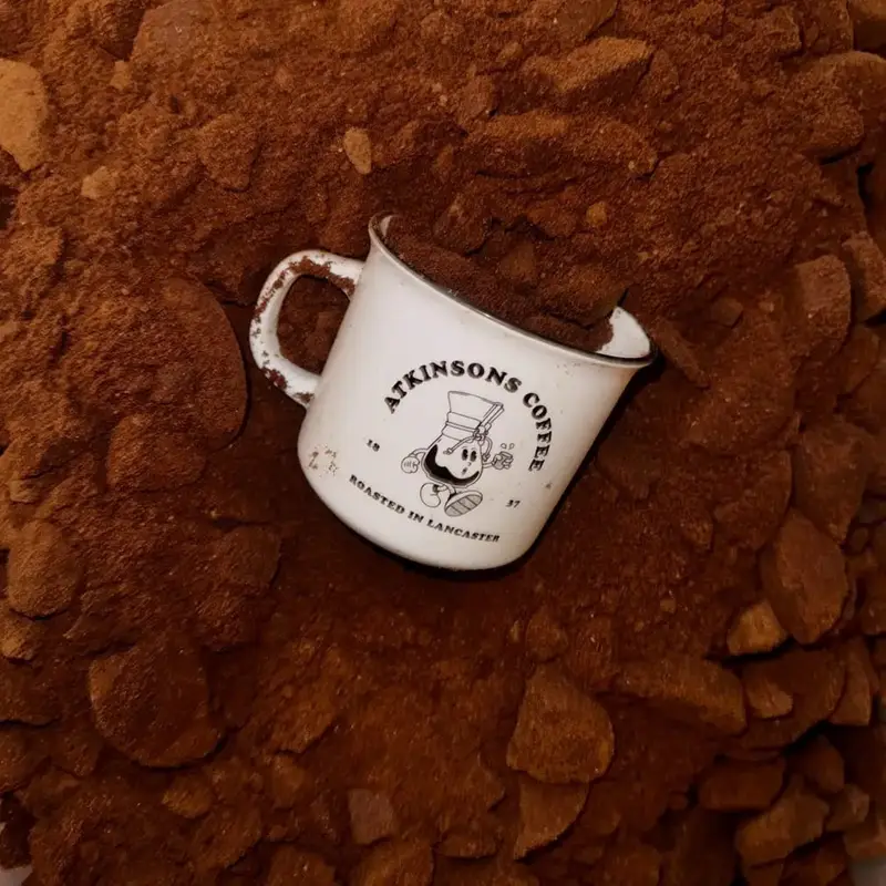 An Atkinsons Coffee enamel mug nestled in a bed of freshly ground coffee