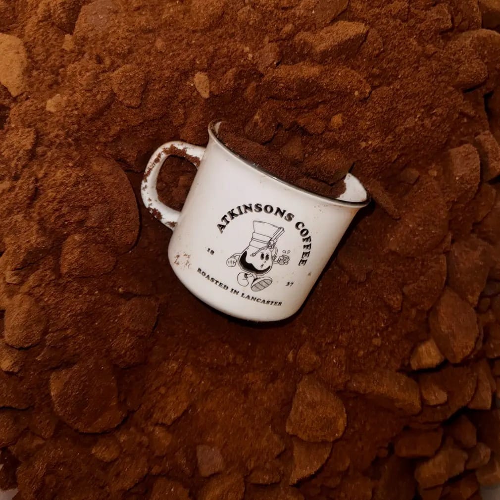 An Atkinsons Coffee enamel mug nestled in a bed of freshly ground coffee